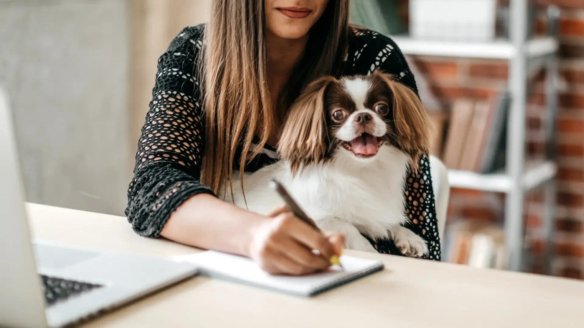 A woman sits at a table with a dog on her lap, writing something on her notepad next to the laptop